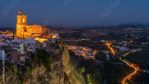 Village of Arcos de la Frontera, Spain, during the blue hour. Iglesia de san pedro is bathed in golden light perched on a cliff with a cobalt blue sky background.
