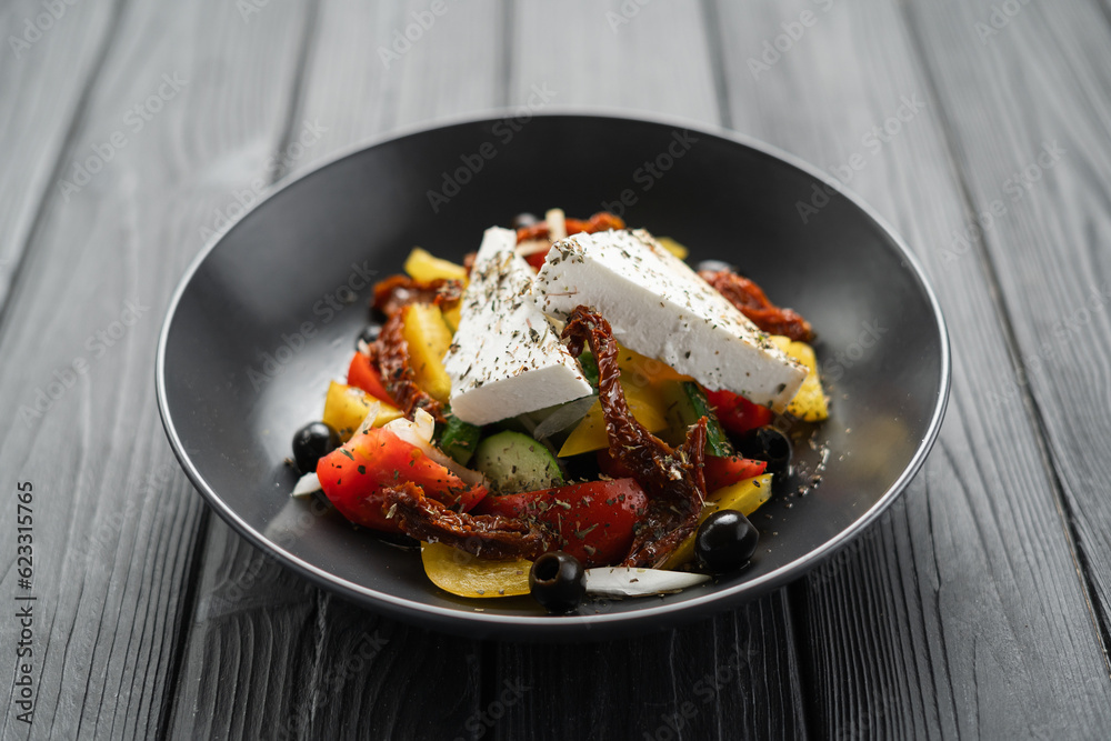 Healthy greek salad of fresh vegetables with tomatoes, bell pepper, cucumbers, olives, feta cheese and sun dried tomatoes in plate on black wooden background. Healthy food, vegetarian dieting Healthy greek salad of fresh vegetables with tomatoes, bell pepper, cucumbers, olives, feta cheese and sun dried tomatoes in plate on black wooden background. Healthy food, vegetarian dieting