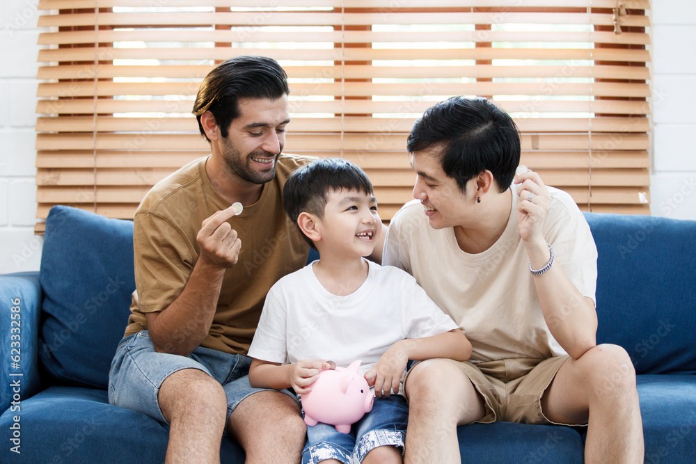 Happy LGBTQ+ gay family with adopted little son stay together in living room and teaching their adopted son about wealth and saving money for future. Diversity in gender and lifestyle concept. Happy LGBTQ+ gay family with adopted little son stay together in living room and teaching their adopted son about wealth and saving money for future. Diversity in gender and lifestyle concept.