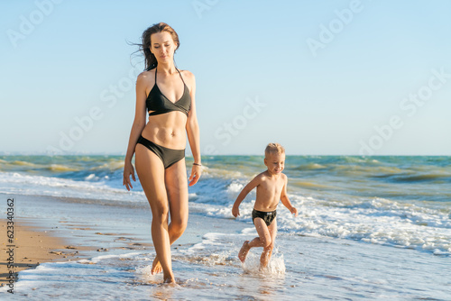 a young woman in a swimsuit with her little son walk along the seashore at sunset