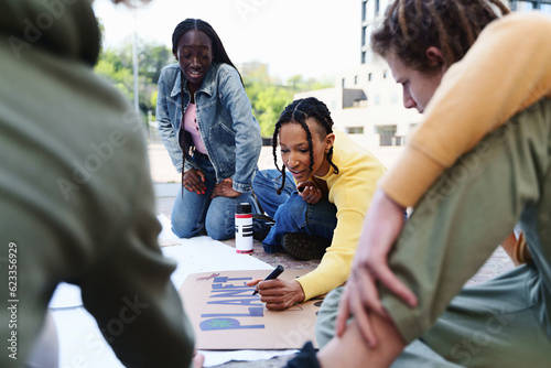 Students Crafting Environmental Protest Signs, Focus on \'Planet\' - African women creating signs for an environmental protest, one writing \'Planet\', a Caucasian male observing in blurred foreground