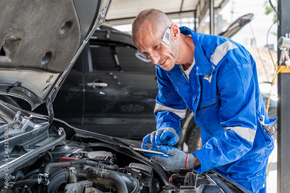 A male car mechanic using his computer tabet to check on the car's engine while working its engine at the car repair garage