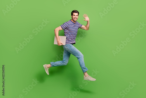 Full length photo of friendly funky man dressed striped t-shirt jumping high holding device waving arm isolated green color background