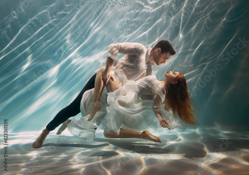 Real People art photo. Happy couple in love swim underwater, woman muse inspires male writer poet creator. Nymph girl dancing with guy at bottom sea under water. Red hair white long silk dress float