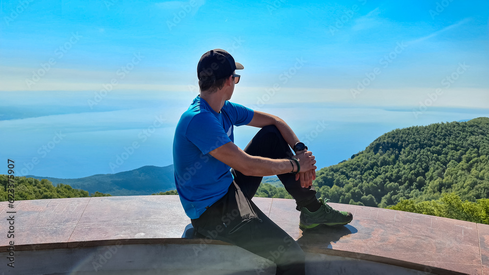 A man sitting on top of Vojak and enjoying the panoramic view on Mediterranean Sea. The mountain is overgrown with lush green plants. Few islands in the back. Early morning hiking by the sea. Remedy A man sitting on top of Vojak and enjoying the panoramic view on Mediterranean Sea. The mountain is overgrown with lush green plants. Few islands in the back. Early morning hiking by the sea. Remedy