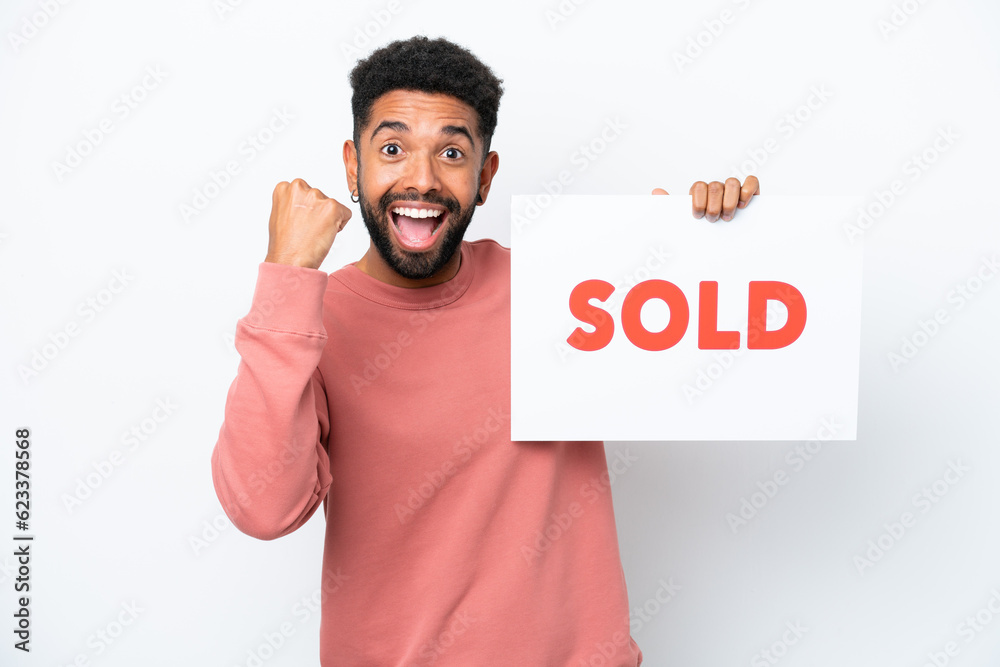 Young Brazilian man isolated on white background holding a placard with text SOLD and celebrating a victory Young Brazilian man isolated on white background holding a placard with text SOLD and celebrating a victory