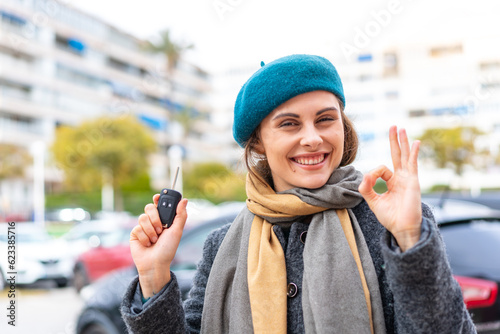 Brunette woman holding car keys at outdoors showing ok sign with fingers