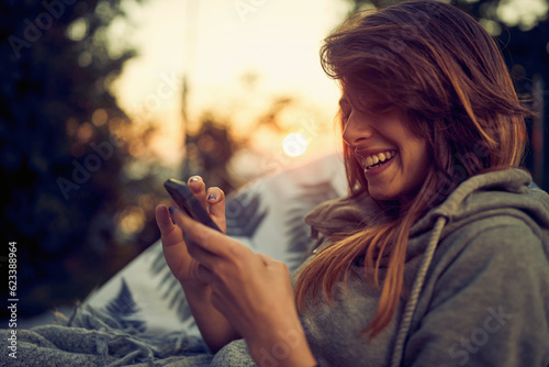 Close up portarit shot of beautiful joyful young woman using smartphone, enjoying her time outdoors, sitting in cozy chair. photo