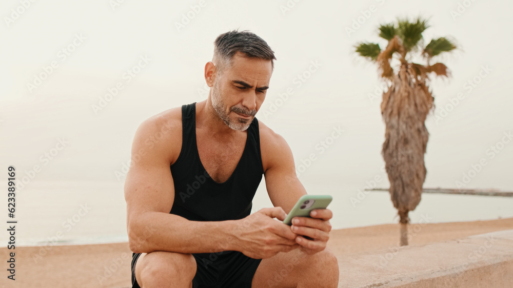 Middle-aged muscular man in sportswear uses smartphone while sitting on promenade on sea background