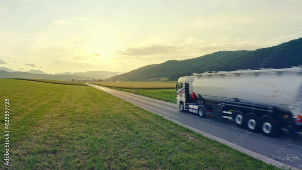 White tanker truck traveling on sunlit highway road aerial shot. Lorry transporting freight while riding through green field with mountains in background