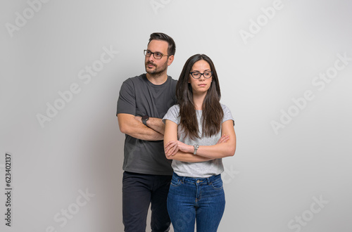 Thoughtful young couple with arms crossed standing confidently against isolated background. Contemplative man and woman wearing eyeglasses and casuals looking away seriously