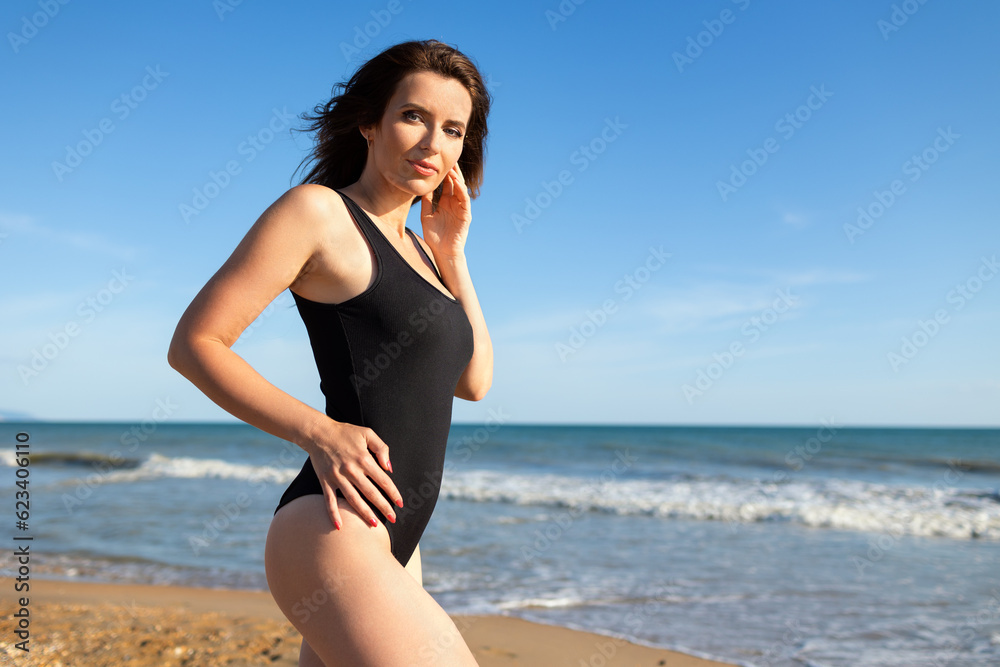 Beautiful brunette woman in a black bodysuit is resting in the summer on a sandy seashore under a blue sky Beautiful brunette woman in a black bodysuit is resting in the summer on a sandy seashore under a blue sky