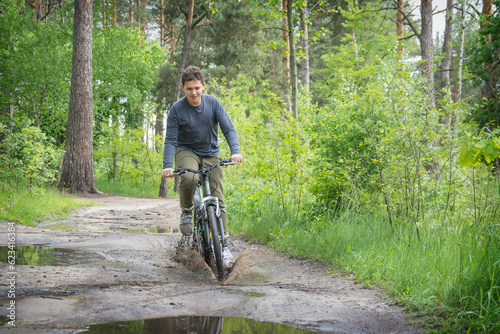 In the summer in the forest, a boy rides a bicycle through a puddle, splashing in different directions.