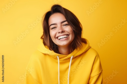 Medium shot portrait photography of a grinning girl in her 20s wearing a comfortable hoodie against a bright yellow background. With generative AI technology © Markus Schröder