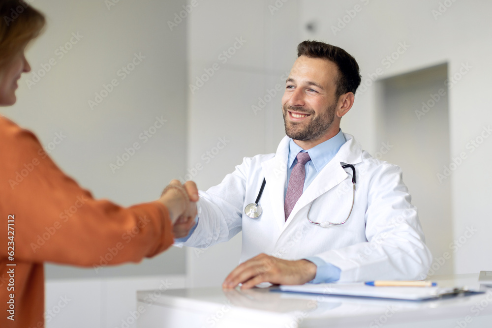 Cheerful male GP shaking hand of female patient at consultation in clinic. Man physician handshaking woman client, closing health insurance deal Cheerful male GP shaking hand of female patient at consultation in clinic. Man physician handshaking woman client, closing health insurance deal