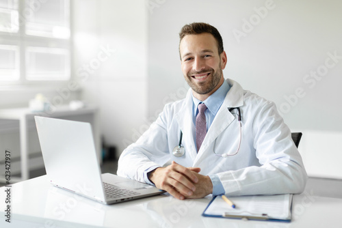 Portrait of cheerful man doctor in workwear working in modern clinic, sitting at workdesk with laptop and medical chart smiling at camera
