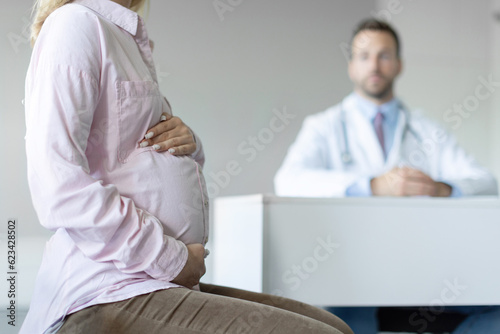 Closeup of woman expecting baby having appointment with doctor in clinic prenatal healthcare center, focus on lady holding hands on baby bump in doc office