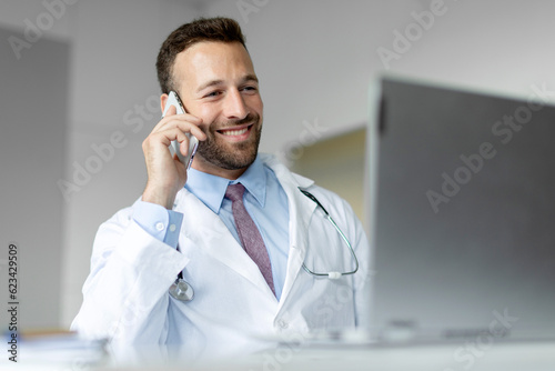 Cheerful male doctor talking on cellphone and using laptop, therapist having mobile conversation with patient, giving recommendations online photo