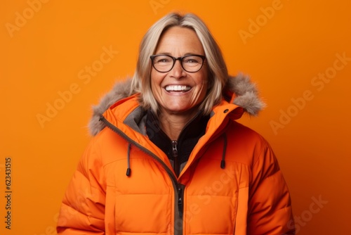 Lifestyle portrait photography of a happy mature woman wearing a warm parka against a tangerine orange background. With generative AI technology