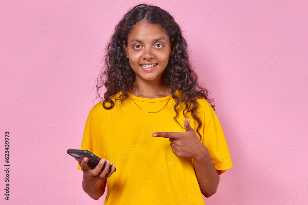 Young happy casual Indian woman teen with black curly hair points finger at mobile phone and smiles looks at screen to promote store selling smartphones stands on isolated pink background. Young happy casual Indian woman teen with black curly hair points finger at mobile phone and smiles looks at screen to promote store selling smartphones stands on isolated pink background.