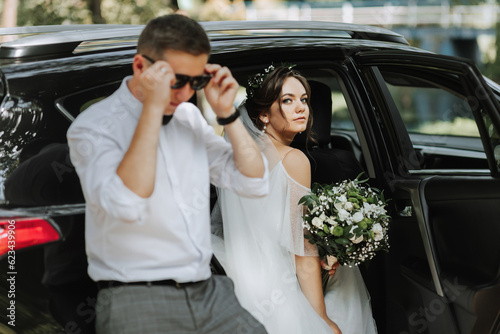 Wedding portrait, photo of a stylish groom in a white shirt and bow tie and a brunette bride with a bouquet of flowers near a black car.