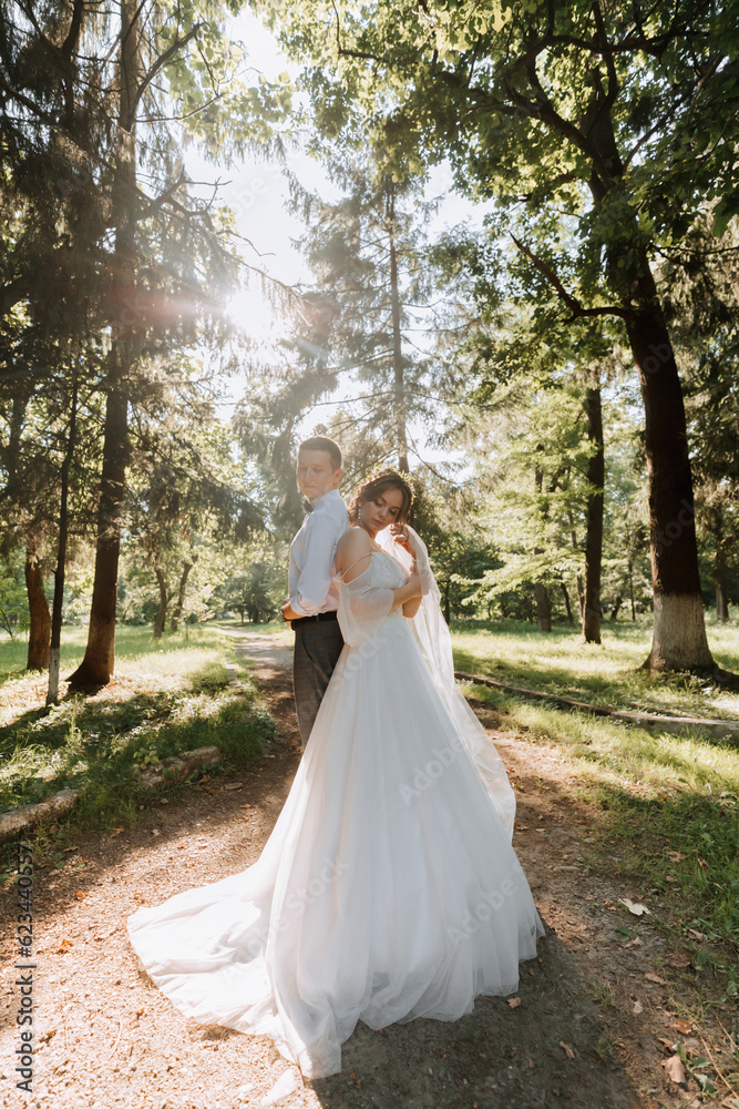 Fashionable groom and cute bride in white dress with tiara of fresh flowers, hugging, laughing in park, garden, forest outdoors. Wedding photography, portrait of smiling newlyweds. Wide angle