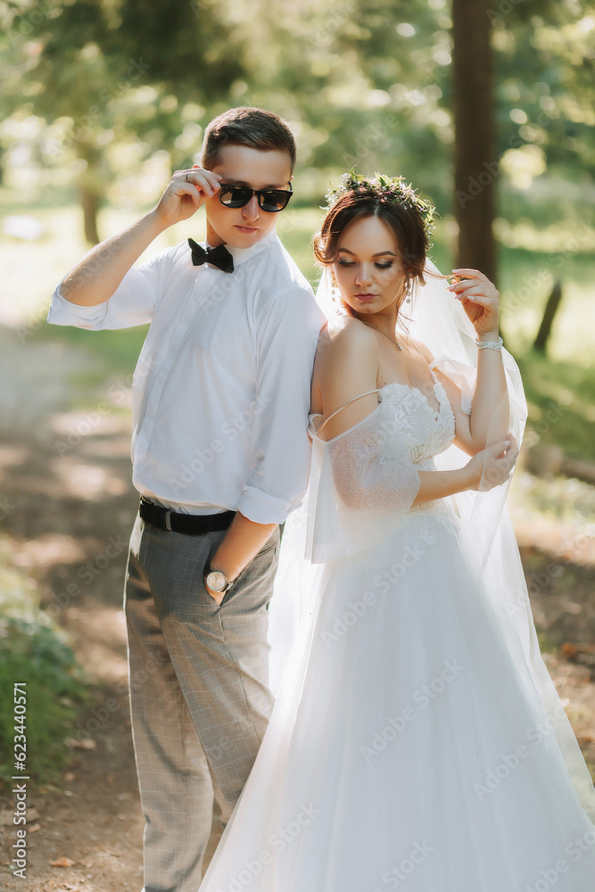 Fashionable groom and cute bride in white dress with tiara of fresh flowers, hugging, laughing in park, garden, forest outdoors. Wedding photography, portrait of smiling newlyweds. Fashionable groom and cute bride in white dress with tiara of fresh flowers, hugging, laughing in park, garden, forest outdoors. Wedding photography, portrait of smiling newlyweds.