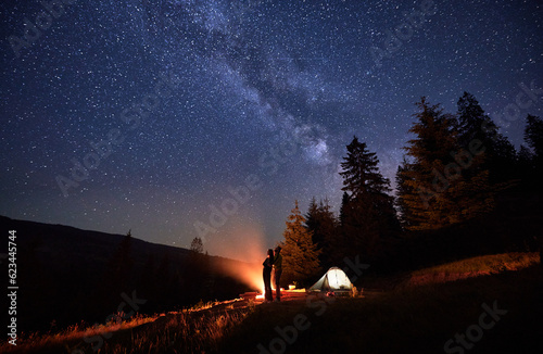 Overnight camping expedition  under shimmering stars. Man and woman intrepid hikers near bonfire and tent pitched near woods  under resplendent night sky with Chumack Path as their celestial guide.