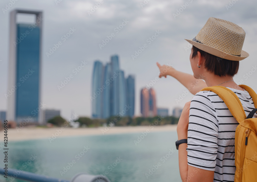 Fototapeta premium travel to the United Arab Emirates, Happy young asian female traveler with backpack and hat view to cityscape of downtown Abu Dhabi. Vacation and tourist destination concept.