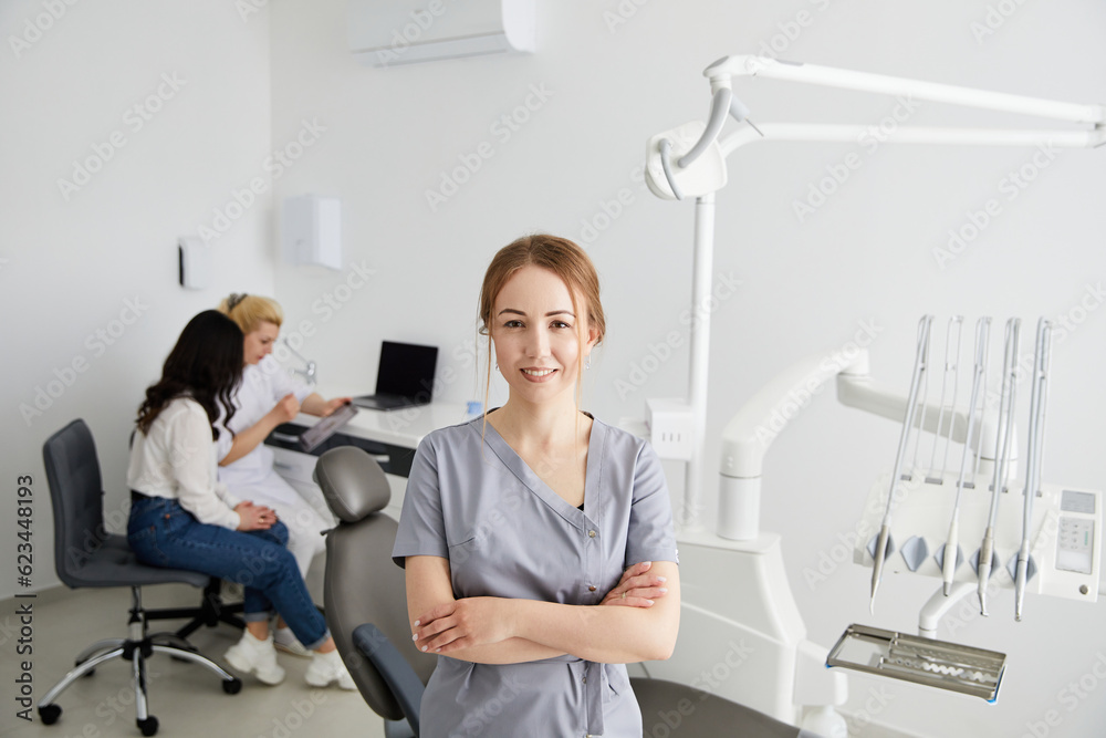 A young pretty female dentist stands in the middle of the office, and in the background a colleague is advising patients.