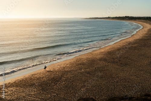 Paisaje de playa solitaria con figura de surfista andando por la playa solo con tabla de surf