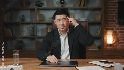 Front view of stressed company owner looking at camera and twisting finger near temple. Worried asian man sitting at office desk with closed laptop and blaming stupid colleague for insane plan.