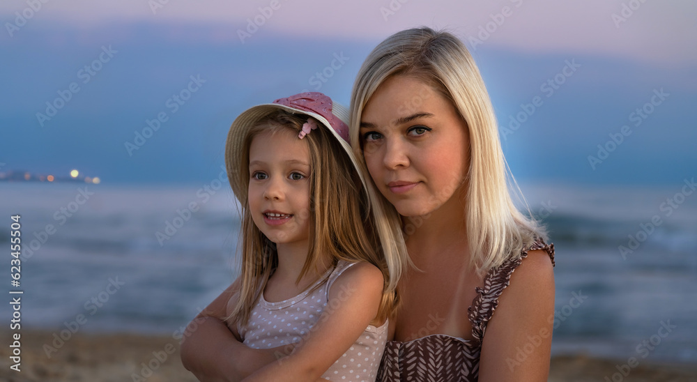 Portrait of mother and daughter on beach of sea or ocean in evening at sunset. Summer vacation, travel, holiday. People outdoors on seascape