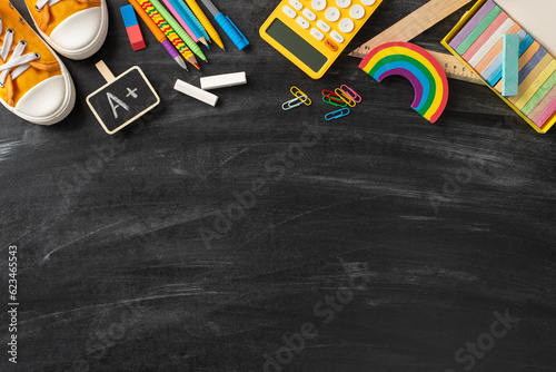 Tools for artistic exploration. Top view of colored pencils, pens, chalk set, A+ reward sticker, ruler, clips, calculator, plasticine, sporty sneakers on a chalkboard background for custom text