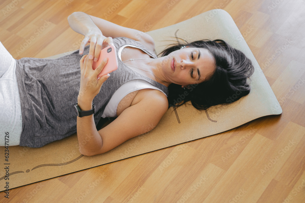 Latin woman lying on a yoga mat at home checking her mobile phone. top view