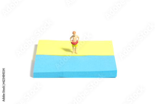 Creative miniature people toy figure photography. Sticky notes installation. Young boy, kids, with rubber ring tube standing at beach shore. Isolated on white background