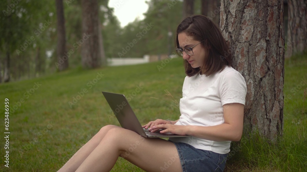 Young woman with laptop in nature. A girl with glasses sits under a tree and types on a laptop. Freelancer woman. Closeup. Young woman with laptop in nature. A girl with glasses sits under a tree and types on a laptop. Freelancer woman. Closeup.