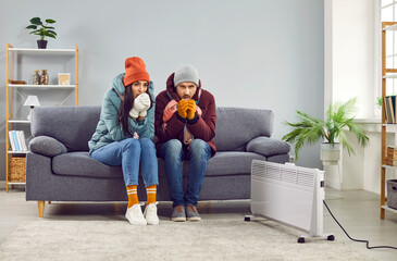 Full length photo of a young frozen family couple man and woman sitting on sofa in the living room in winter outerwear and in hats at home warming their hands. Heating problems concept.