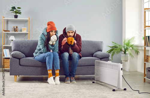 Fototapete Full length photo of a young frozen family couple man and woman sitting on sofa in the living room in winter outerwear and in hats at home warming their hands