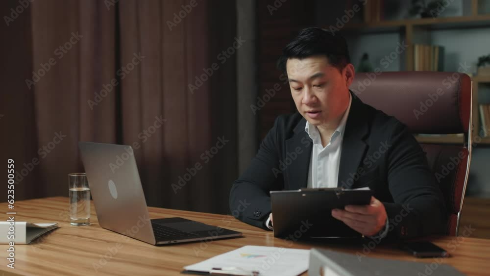 Serious company ceo in business clothes talking to employee over video call while staying in contemporary office space. Focused asian entrepreneur discussing company issues using portable computer. Serious company ceo in business clothes talking to employee over video call while staying in contemporary office space. Focused asian entrepreneur discussing company issues using portable computer.