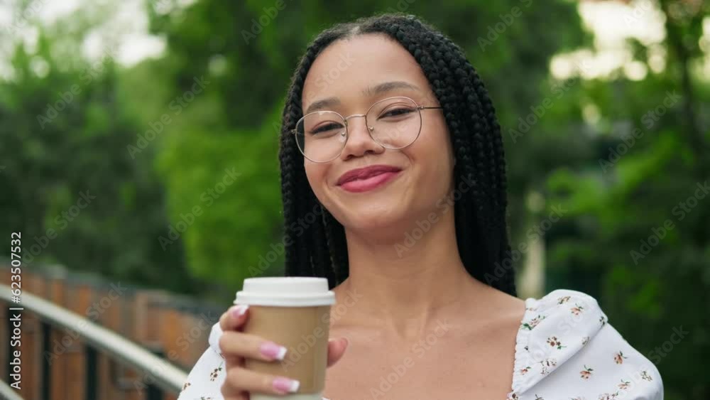Portrait of an attractive young African woman with glasses, looking at the camera, smiling and drinking coffee in the park on a sunny summer day. Close up Portrait of an attractive young African woman with glasses, looking at the camera, smiling and drinking coffee in the park on a sunny summer day. Close up