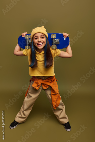 Full length of smiling preteen child with dyed hair wearing yellow hat and urban outfit holding skateboard and looking at camera on khaki background, girl with cool street style look