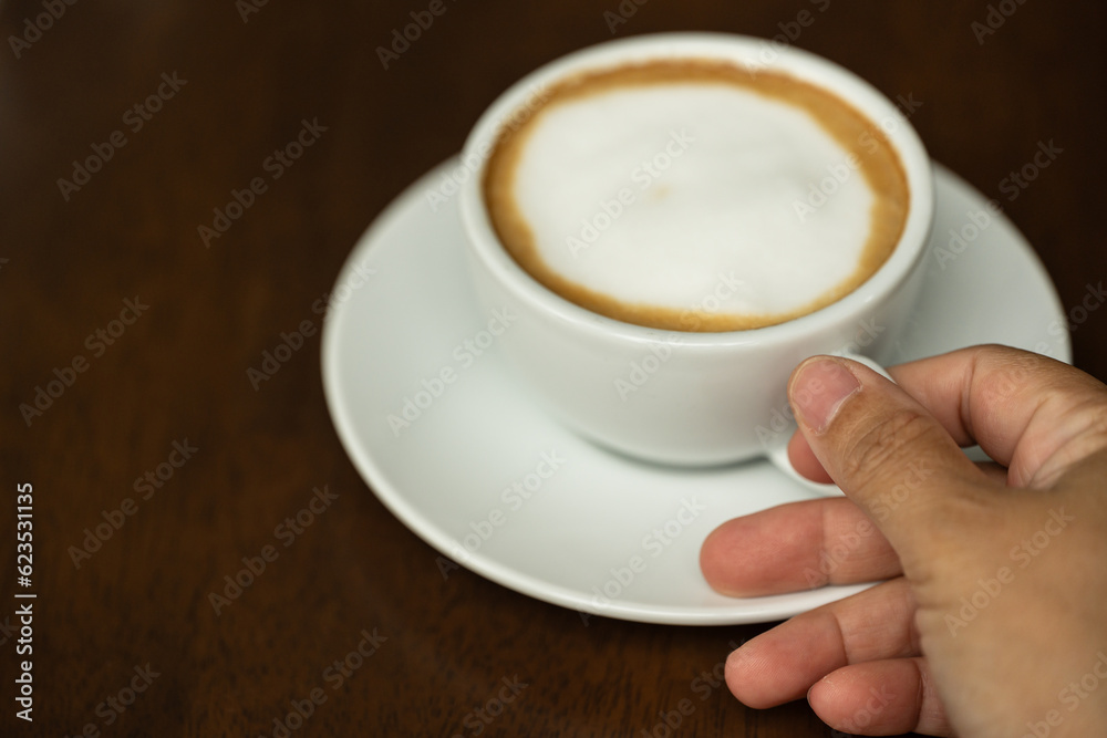 Closeup hand holding white cup of hot coffee latte on wood background in restaurant.Best of menu in the coffee shop.Soft focus. Closeup hand holding white cup of hot coffee latte on wood background in restaurant.Best of menu in the coffee shop.Soft focus.