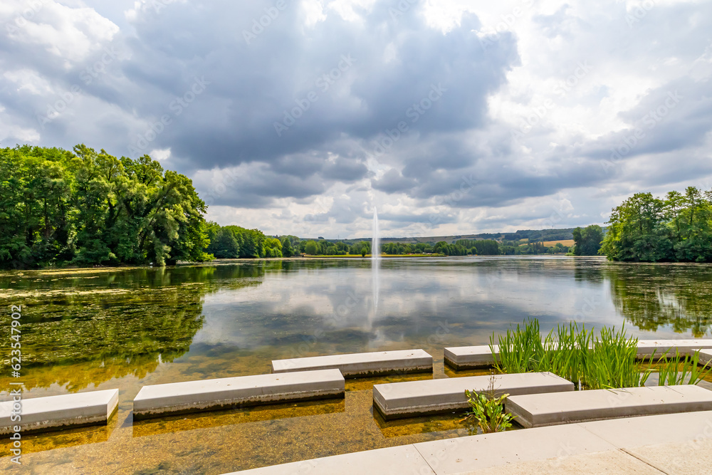 Fototapeta premium Concrete platforms on shore of Lake Echternach with waterjet gushing from a fountain, green leafy trees in background, reflection on water surface, panoramic landscape, summer cloudy day in Luxembourg