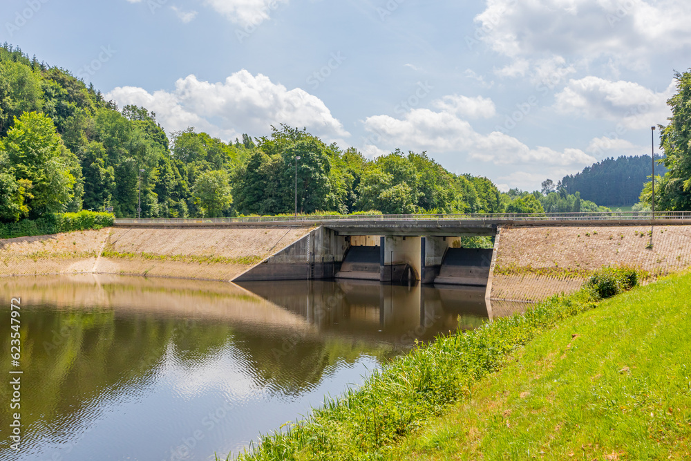 Staumauer Bitburger dam on river Prum, reflection on water surface, vehicular bridge, green leafy trees in background, Stausee Bitburg reservoir on a sunny spring day in Germany Staumauer Bitburger dam on river Prum, reflection on water surface, vehicular bridge, green leafy trees in background, Stausee Bitburg reservoir on a sunny spring day in Germany
