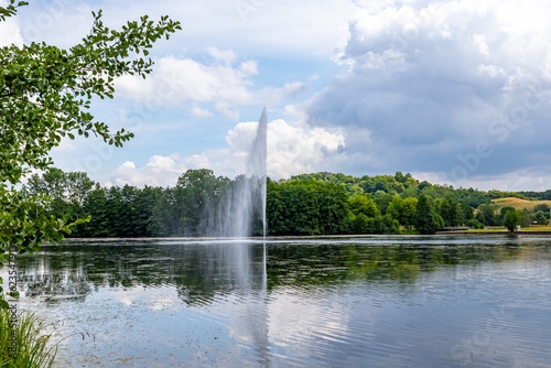 Echternach lake with a waterjet and breeze from a fountain, hills and green leafy trees on misty background, reflection on water surface, sunny spring day with abundant clouds in Luxembourg