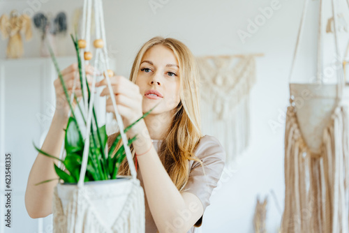 Freelancer woman working on half-finished macrame piece, Blond craft woman holding a potted plant in macrame suspending basket holder. Creative knitting concept. photo