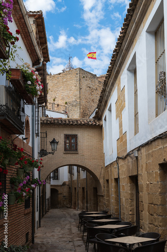 Fototapeta Naklejka Na Ścianę i Meble -  Vista de una callejuela típica con arco y torre con la bandera de España en Baeza, Jaén, Andalucía, España.