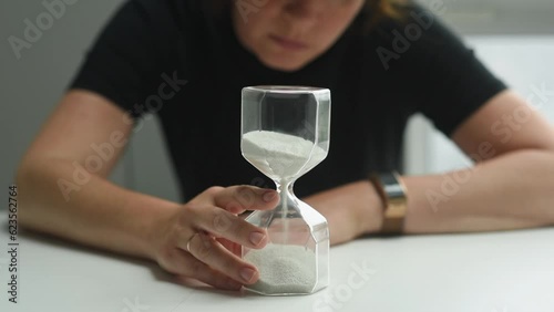 A woman sits at a table and looks at an hourglass, then turns it over - a reversal of time. A woman is stressed that time is running out.