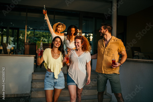 A group of young people exchange enthusiastic greetings, waving and hugging each other, with cocktail bottles in hand going to watch a sunset.